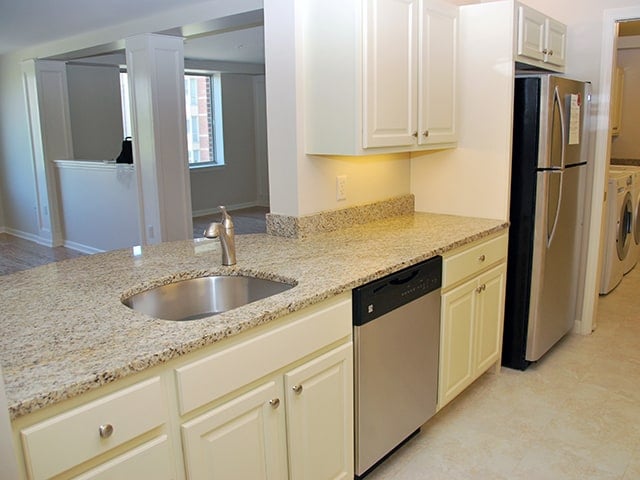 off white cabinets and a brown speckled granite countertop with a stainless steel sink, dishwasher and refrigerator in a Linden Ponds apartment in Hingham, MA